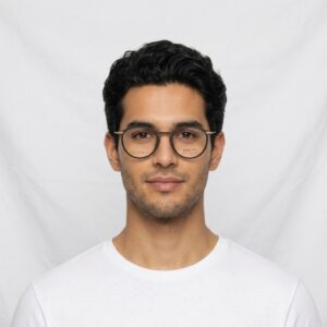Portrait of a man with dark hair and round glasses, wearing a white t-shirt, facing the camera against a white backdrop.