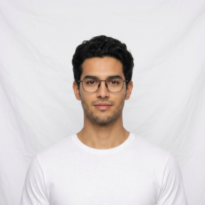 Portrait of a young man with dark hair and round glasses wearing a white T-shirt against a white backdrop in a head-and-shoulders shot.