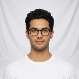 Young man with dark hair and tortoiseshell glasses, wearing a white T-shirt against a plain white backdrop.