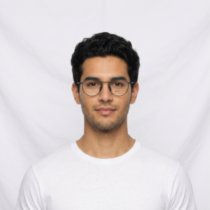 Portrait of a man with dark hair and round glasses, wearing a white t-shirt, against a plain light backdrop.